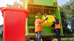 Two men in hi-vis orange jackets, yellow hard hats and gloves tipping a yellow bin into a green waste disposal lorry, red waste bin in the foreground.
