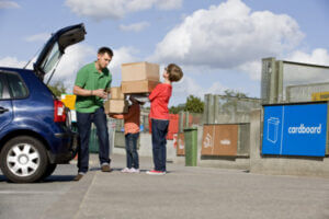 Young light-skinned man lifting cardboard boxes out of his car boot and handing them to his two children who are holding the boxes, they're standing in front of the 'cardboard' recycling sign at a household waste recycling centre.