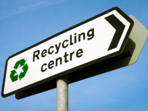 Road sign against a blue sky reading 'Recycling Centre' with black arrow pointing right and the green recycling logo.