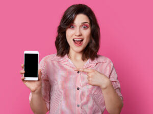 Young light skinned woman with a pink stripy shirt, pink eyeshadow and an excited expression against a bright pink background, she is holding a white smart phone with one hand and pointing to it with the other hand.